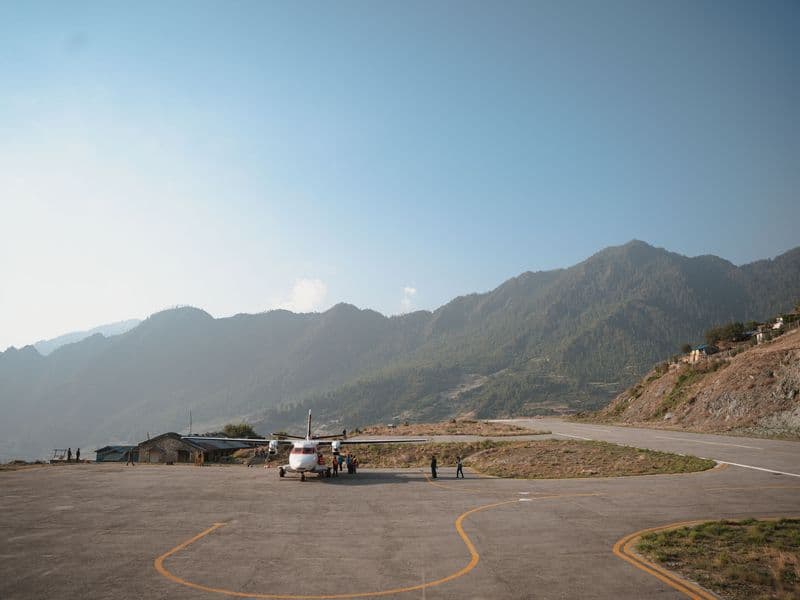 A scenic view of a small aircraft on a mountain airstrip surrounded by lush green hills under a vibrant blue sky.
